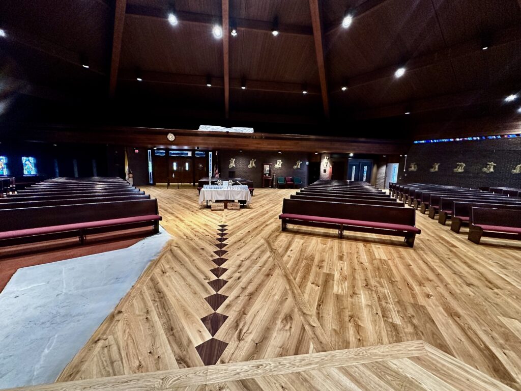 View from the altar toward the church entrance showing wide plank European oak floor with walnut and oak diamond inlays and aligned pews at Saint Thomas More Church